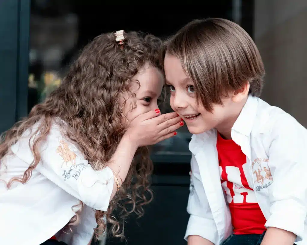 Ein Junge und ein Mädchen. Das Mädchen hält die Hand vor den Mund und flüstert dem Jungen etwas ins Ohr. Der Junge lächelt.