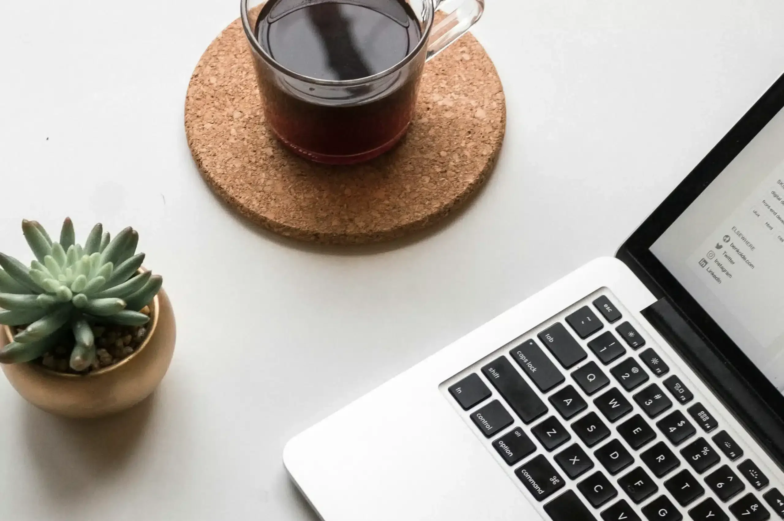 Draufsicht auf einen Schreibtisch mit aufgeklapptem Laptop, iPhone, kleinem Kaktus und einem Kaffee